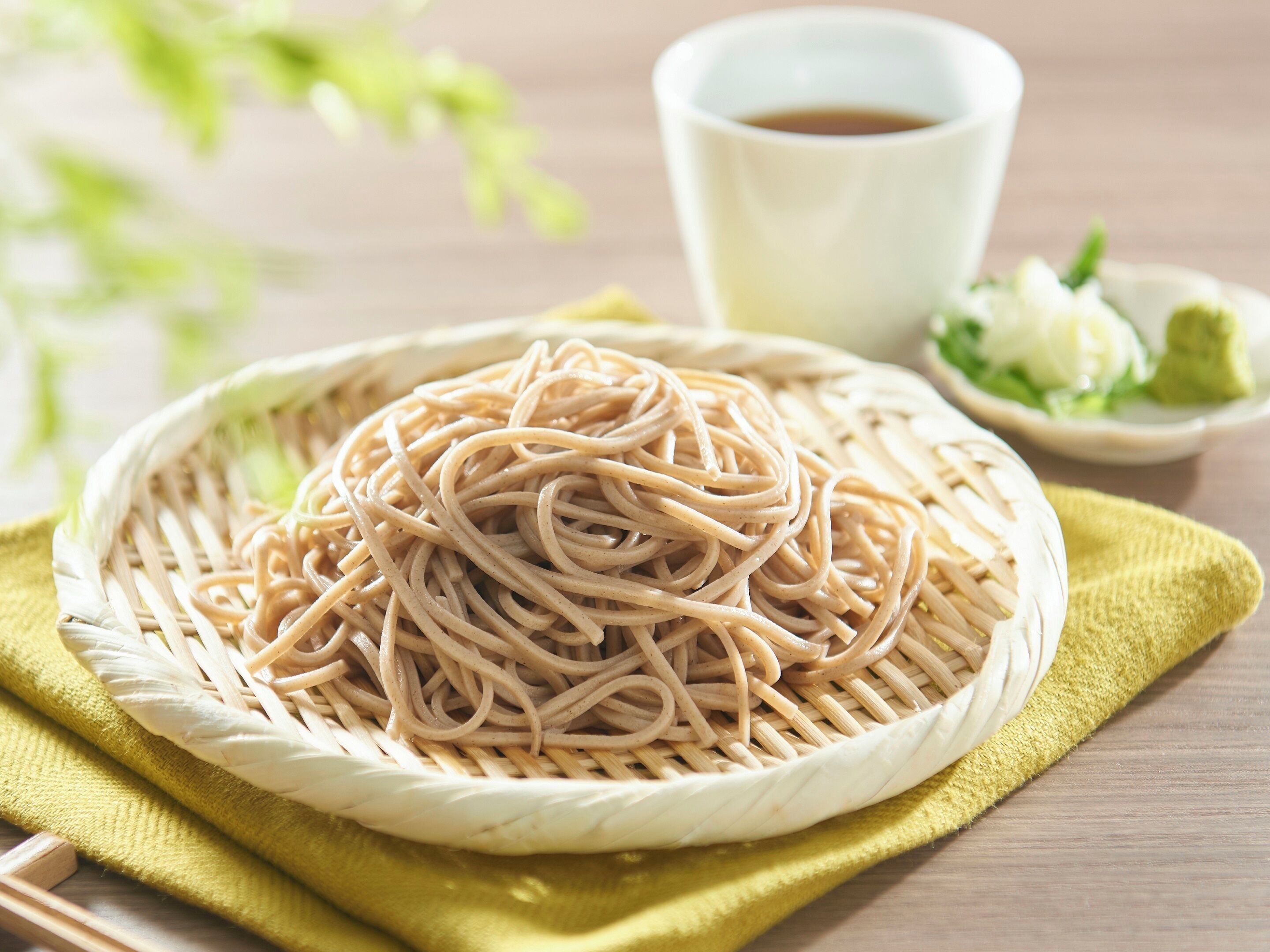 Un panier tressé rempli de nouilles, posé sur une table en bois, accompagné d’une tasse de thé et d’un petit bol de légumes verts.