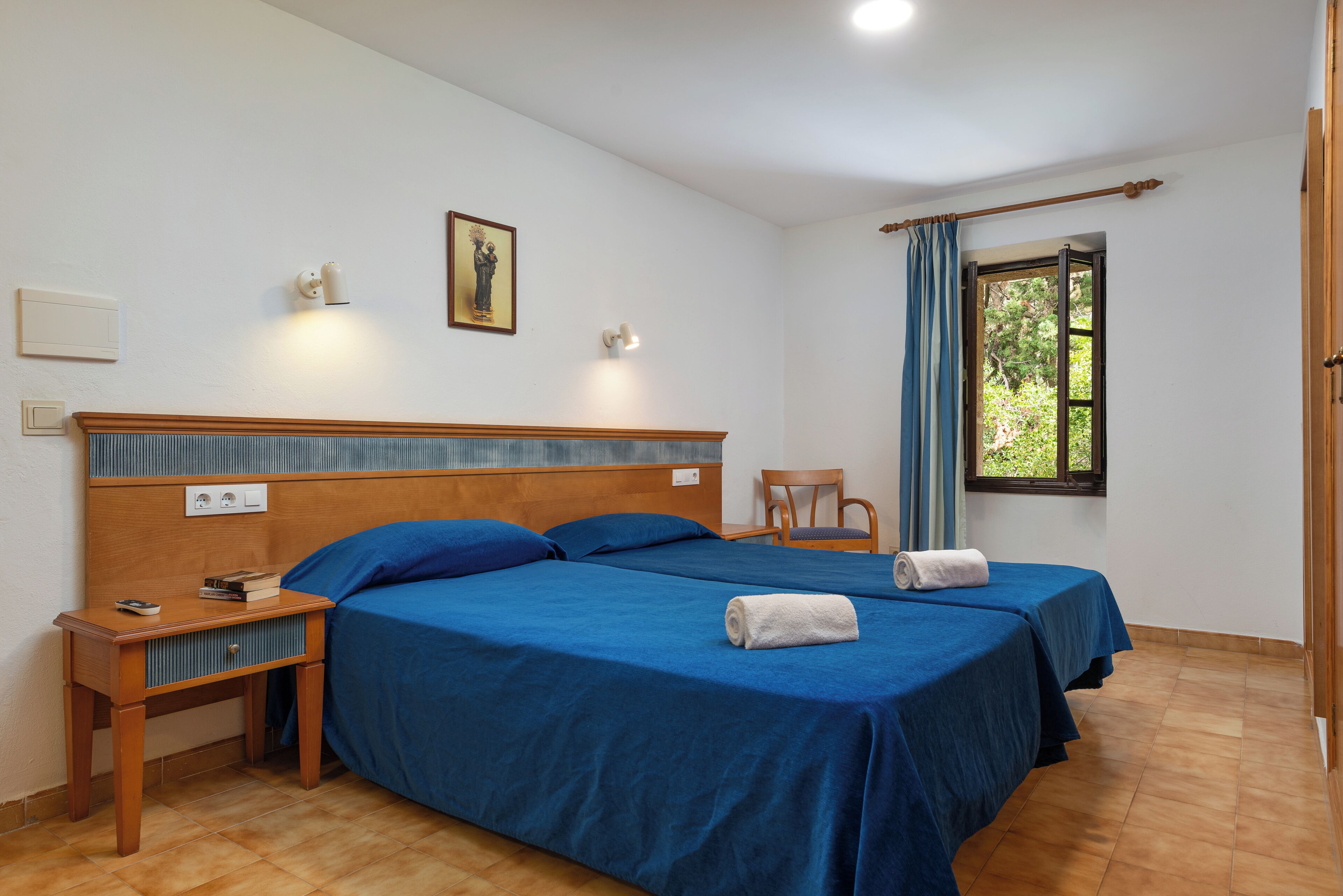 Habitación de hotel con dos camas, una mesita de noche, una silla, una ventana con cortinas y una lámpara fijada a la pared.