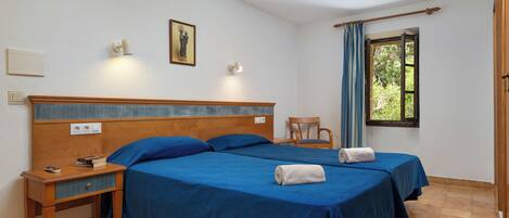 Habitación de hotel con dos camas, una mesita de noche, una silla, una ventana con cortinas y una lámpara fijada a la pared.