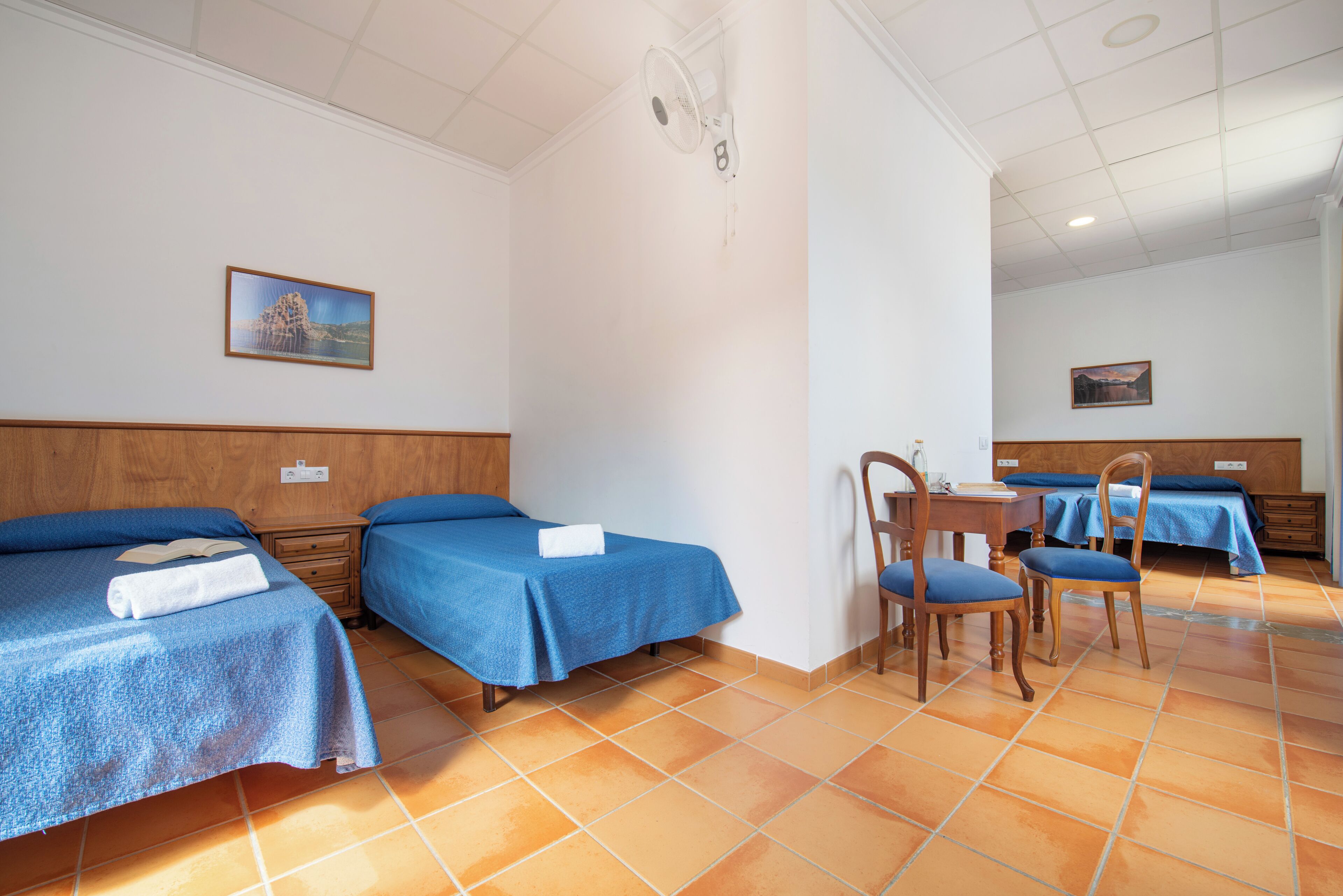 Habitación de hotel con dos camas individuales, una mesita con sillas y un ventilador de techo.