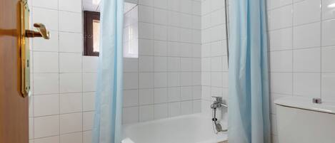 Une salle de bain avec des murs recouverts de carreaux blancs, une baignoire et un rideau de douche.
