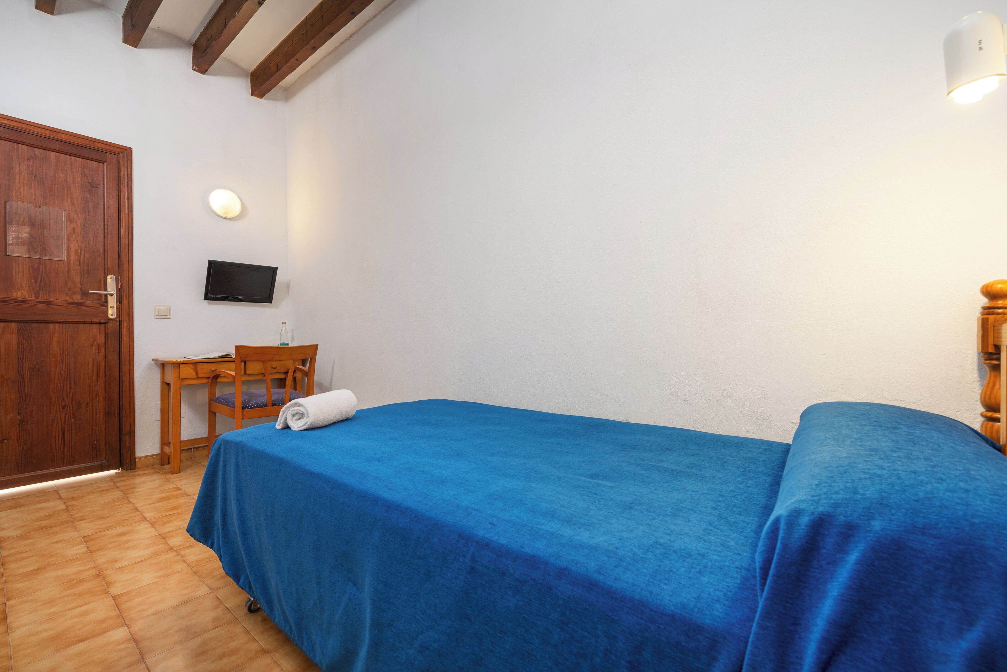 A room with a wooden door, a desk with a chair, a bed with a blue bedspread, a television mounted on the wall, and a small table with a bottle.