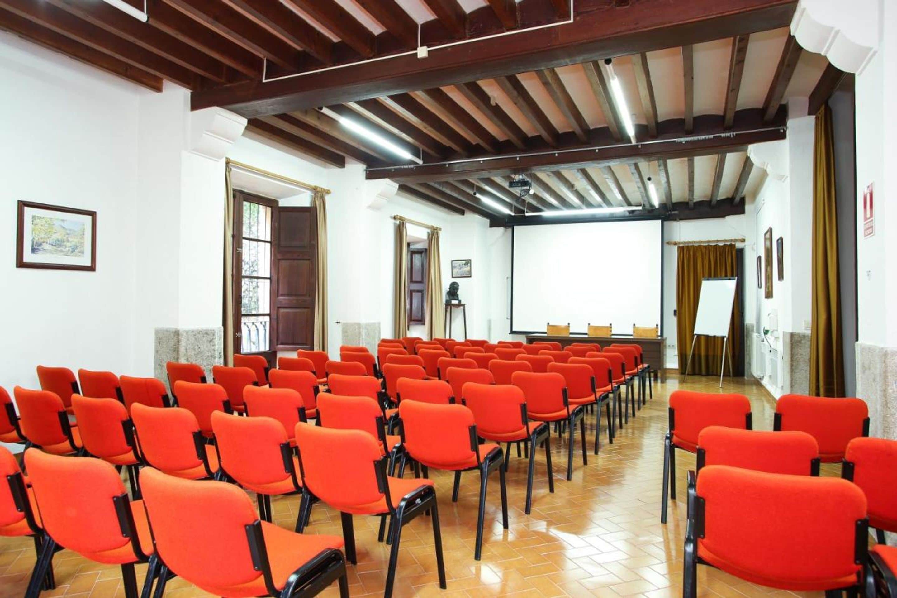 Un salón de conferencias con filas de sillas anaranjadas, vigas de madera en el techo y una pantalla de proyector al frente.