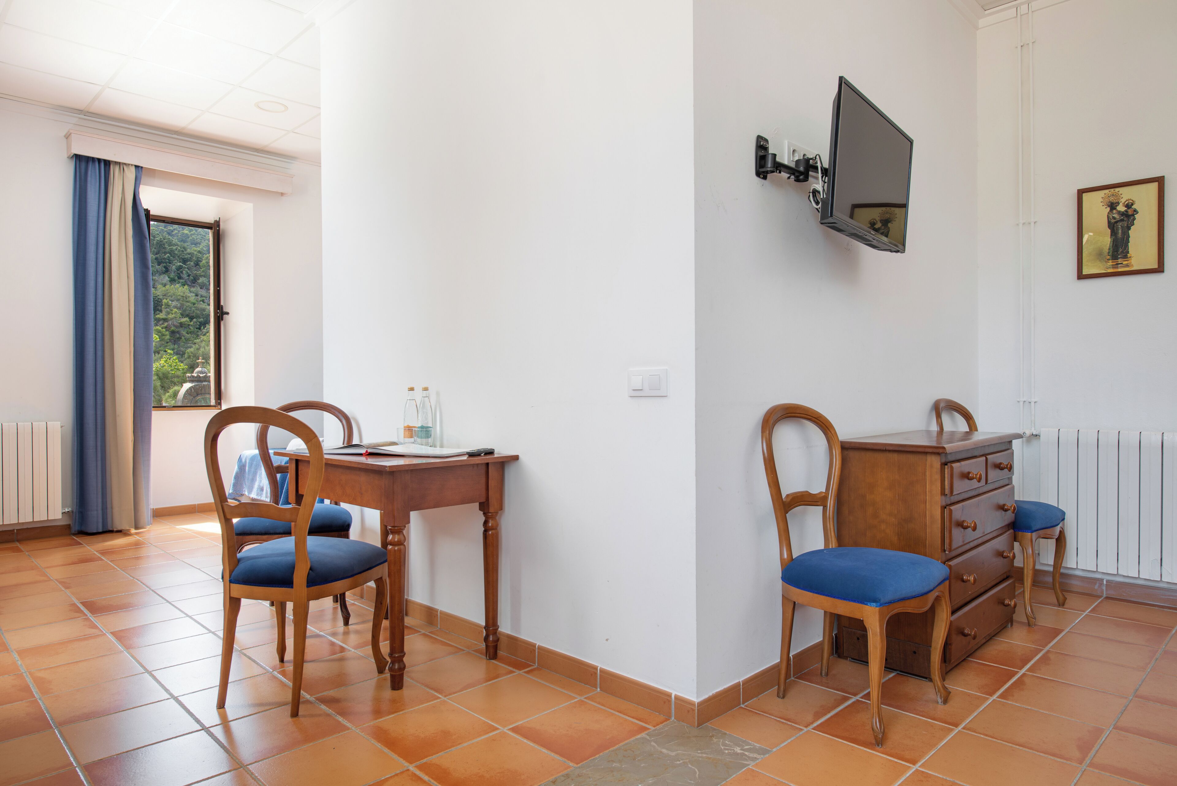 A room with a wooden desk, two chairs, a dresser, a television, and a window with curtains.