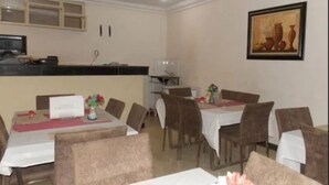 A dining area with tables set for four, a kitchen area, and a framed artwork on the wall. - Gade Place Hotel (Rumuekini)