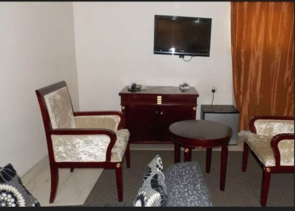 A small, furnished room with two wooden chairs, a small round table, a TV mounted on the wall, and a desk with a telephone.
