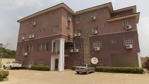 A multi-story building with a symmetrical facade, air conditioning units, and a paved driveway with parked cars. - Gade Place Hotel (Rumuekini)