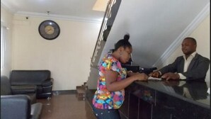 A reception area with a receptionist and a guest. There is a clock on the wall, a staircase, and a reception desk. - Gade Place Hotel (Rumuekini)