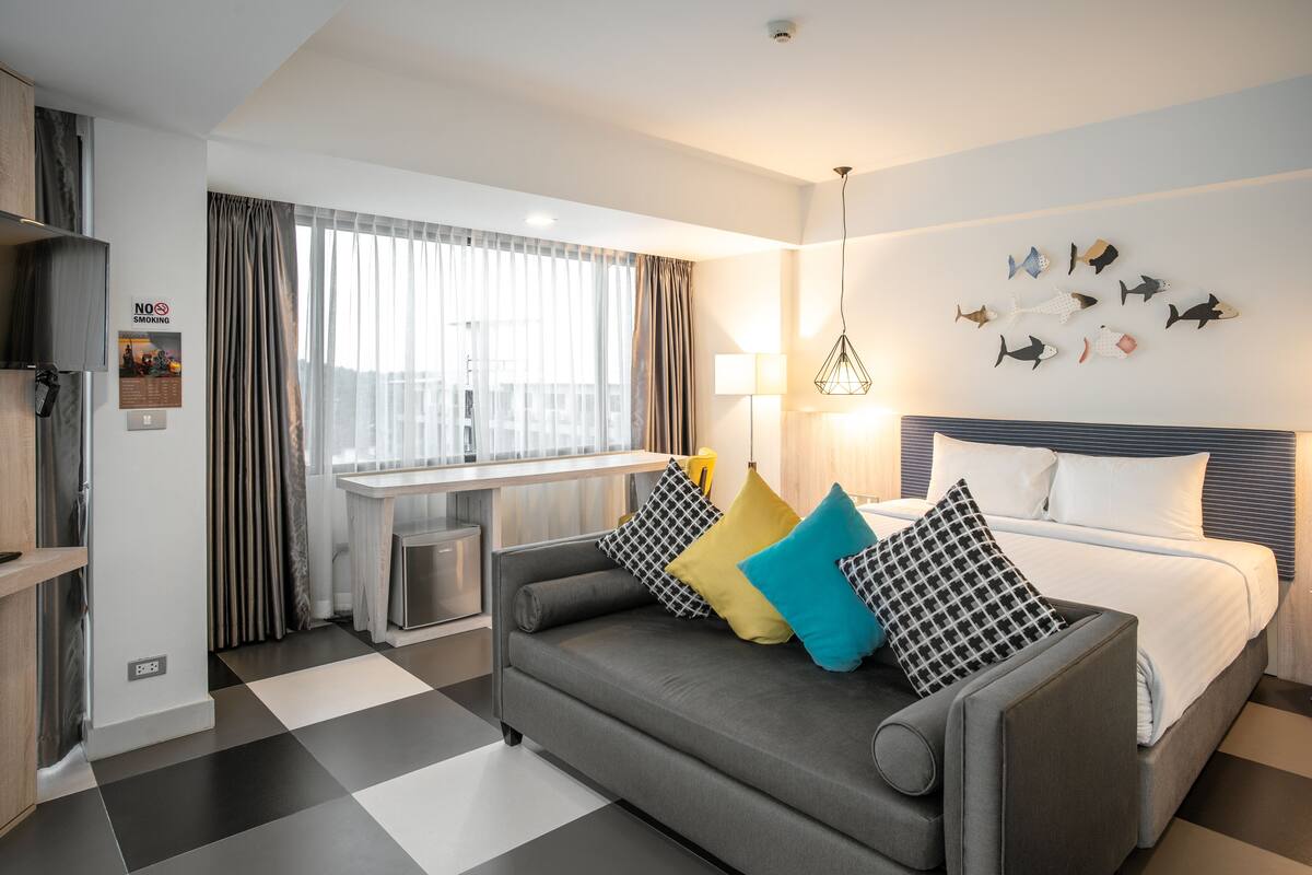 Een moderne hotelkamer met een grijze bank, een bed met wit beddengoed, een visdecoratie aan de muur en een raam met dunne gordijnen.