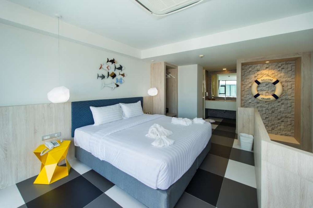 A modern hotel room with a large bed, a unique yellow side table, and a welcome sign on the wall.