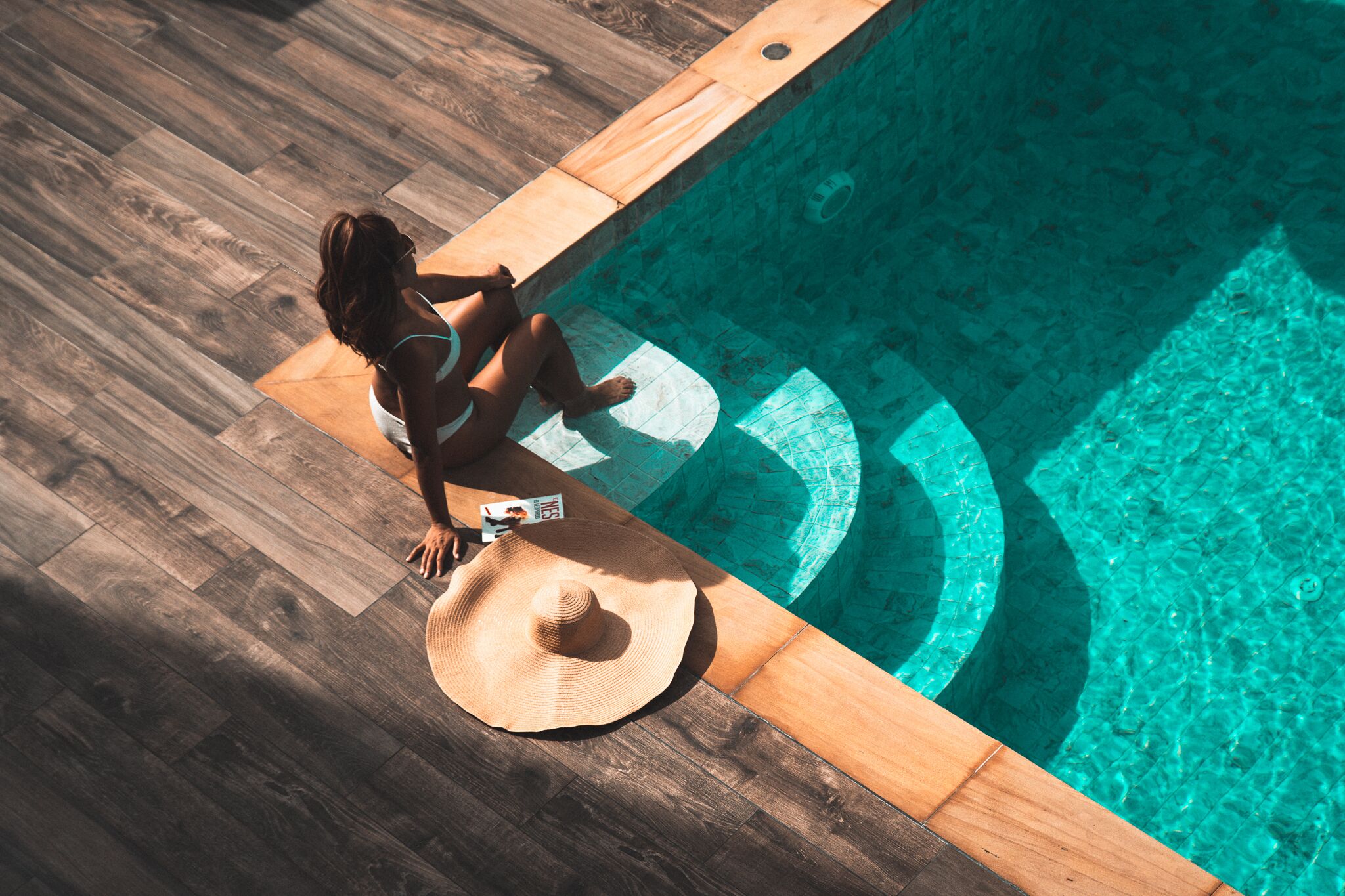A person sitting by a pool with a wooden deck and a hat on the deck.