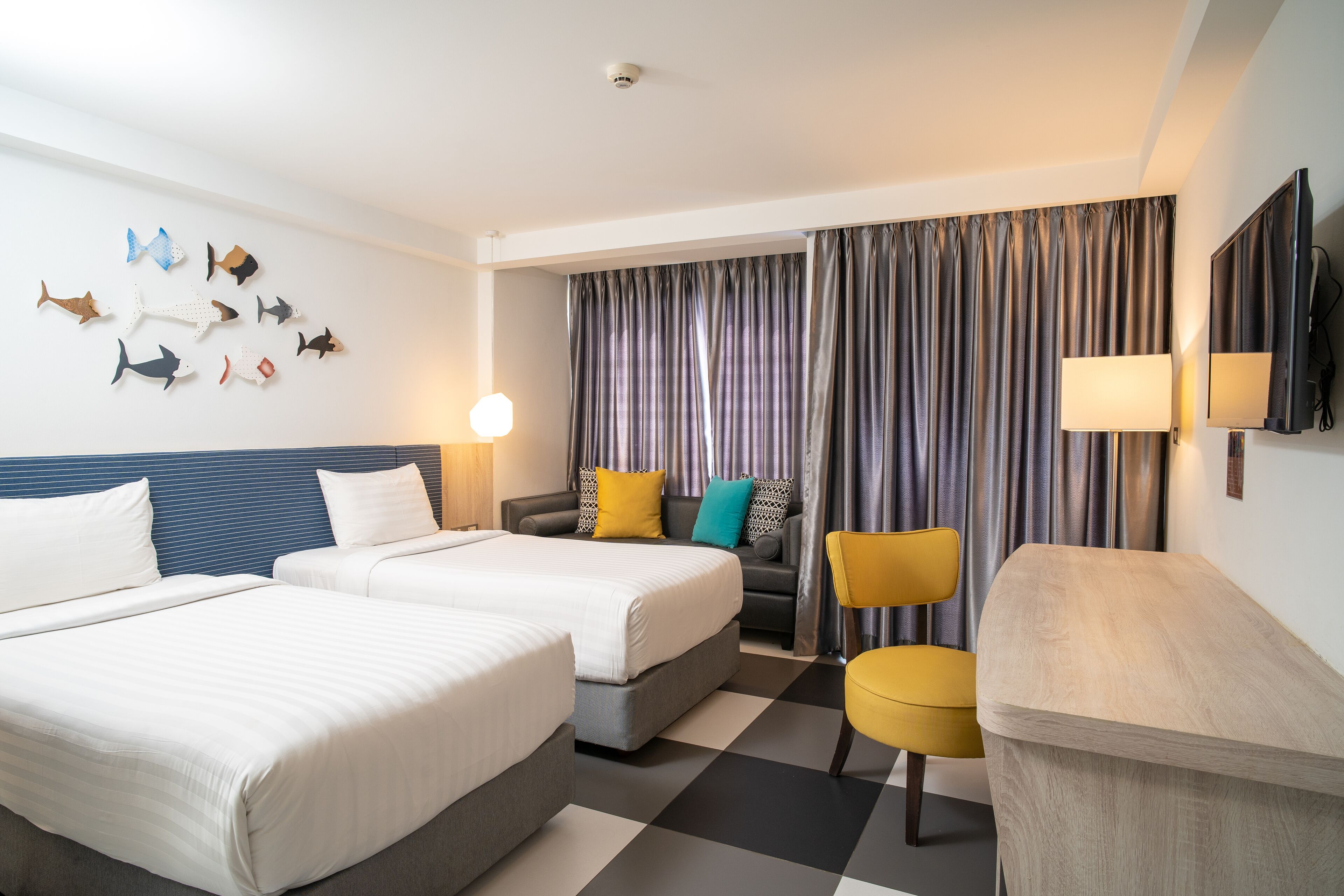 A hotel room with two beds, a sofa, a chair, a flat-screen TV, and a wall-mounted lamp.