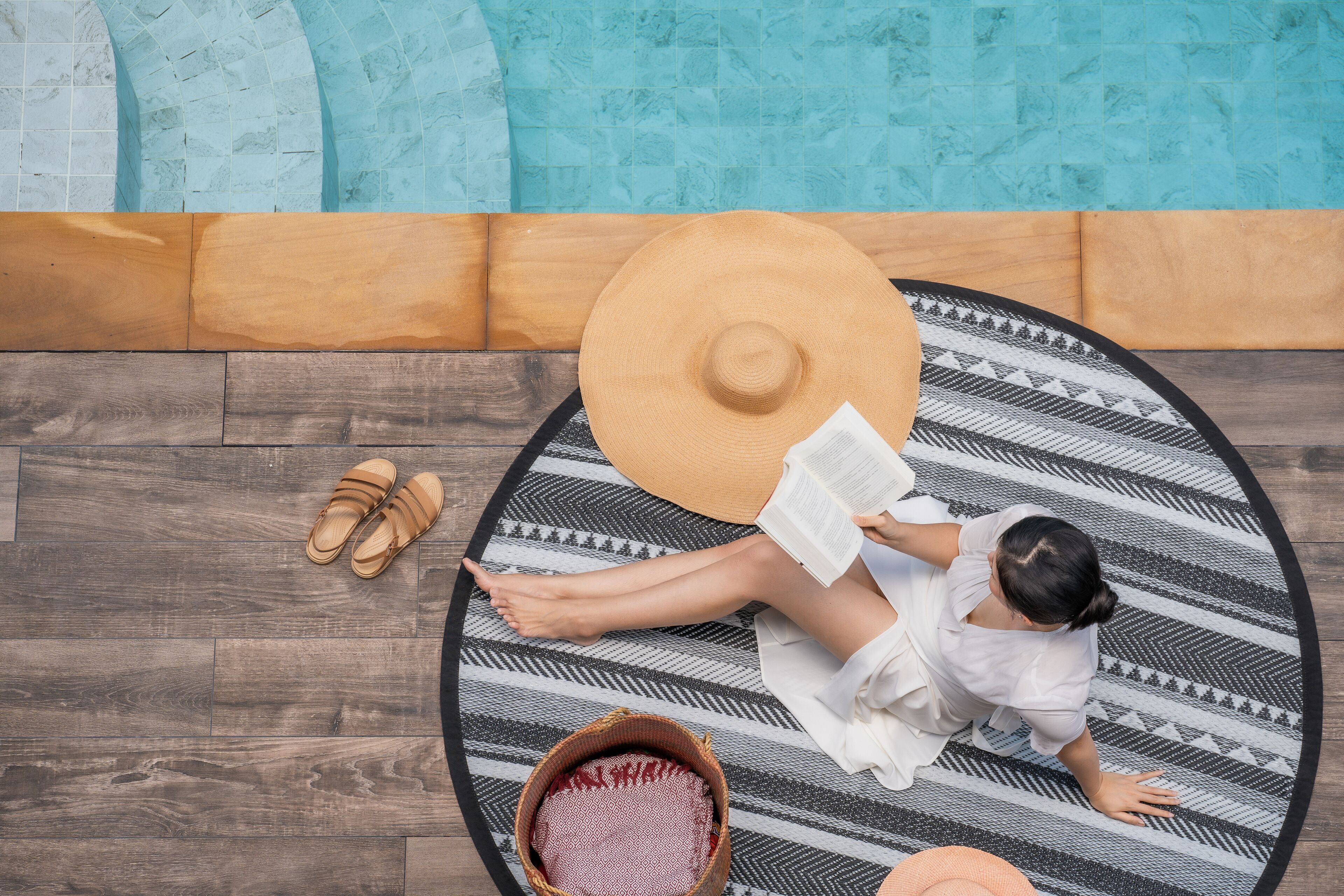 Une personne allongée sur un tapis à motifs près d’une piscine, en train de lire un livre.