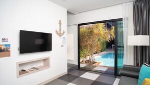 Una habitación de hotel moderna con una televisión de pantalla plana, una puerta corredera de vidrio hacia un área de piscina, un sofá con cojines y un pequeño estante con objetos.