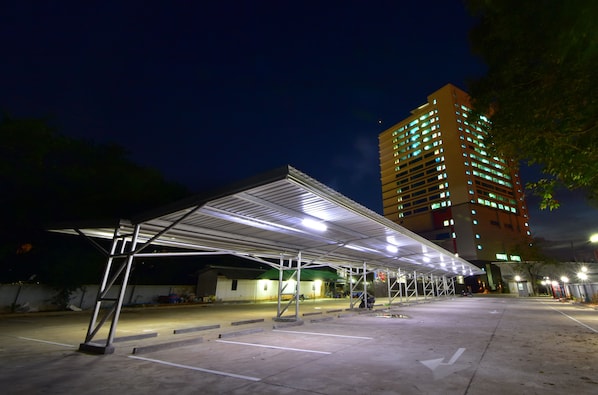 Un espace de stationnement couvert, doté d’une structure de toit en métal et d’un revêtement de sol en béton, jouxtant un immeuble à plusieurs étages dont les fenêtres sont illuminées.