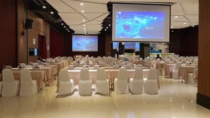Een vergaderzaal met rijen tafels en stoelen, grote schermen waarop presentaties worden getoond, en een podium.