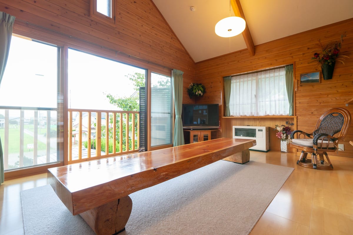 Een ruime woonkamer met een houten bank, een televisie en een groot raam met uitzicht op groen.