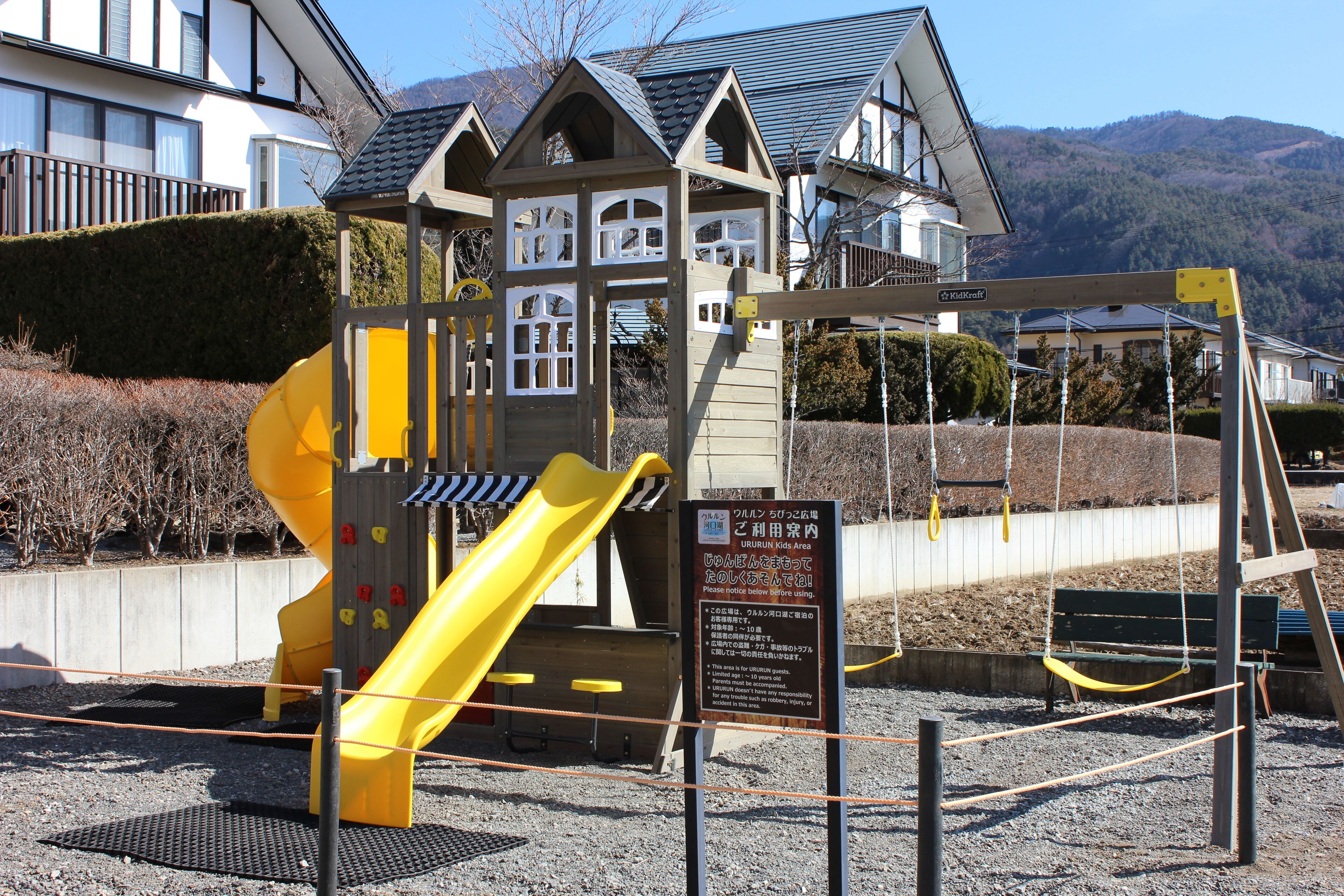 A playground with a yellow slide and a swing set, surrounded by a gravel area and a wooden fence.