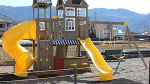 Een houten speelconstructie met een gele glijbaan en klimmuur, met bergen op de achtergrond.