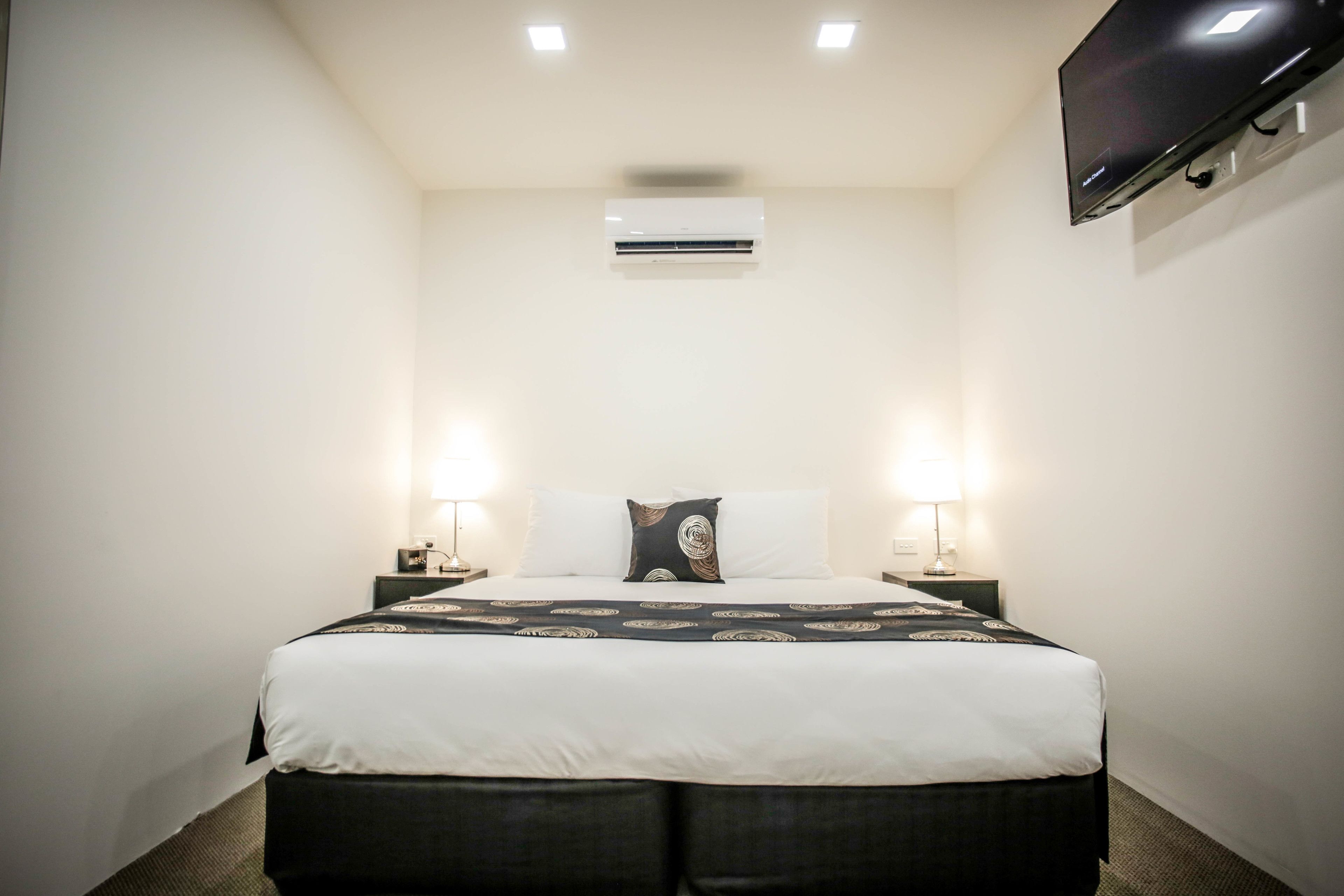 Quarto de hotel com uma cama grande, duas luminárias laterais, uma televisão de tela plana na parede e um aparelho de ar condicionado no teto.