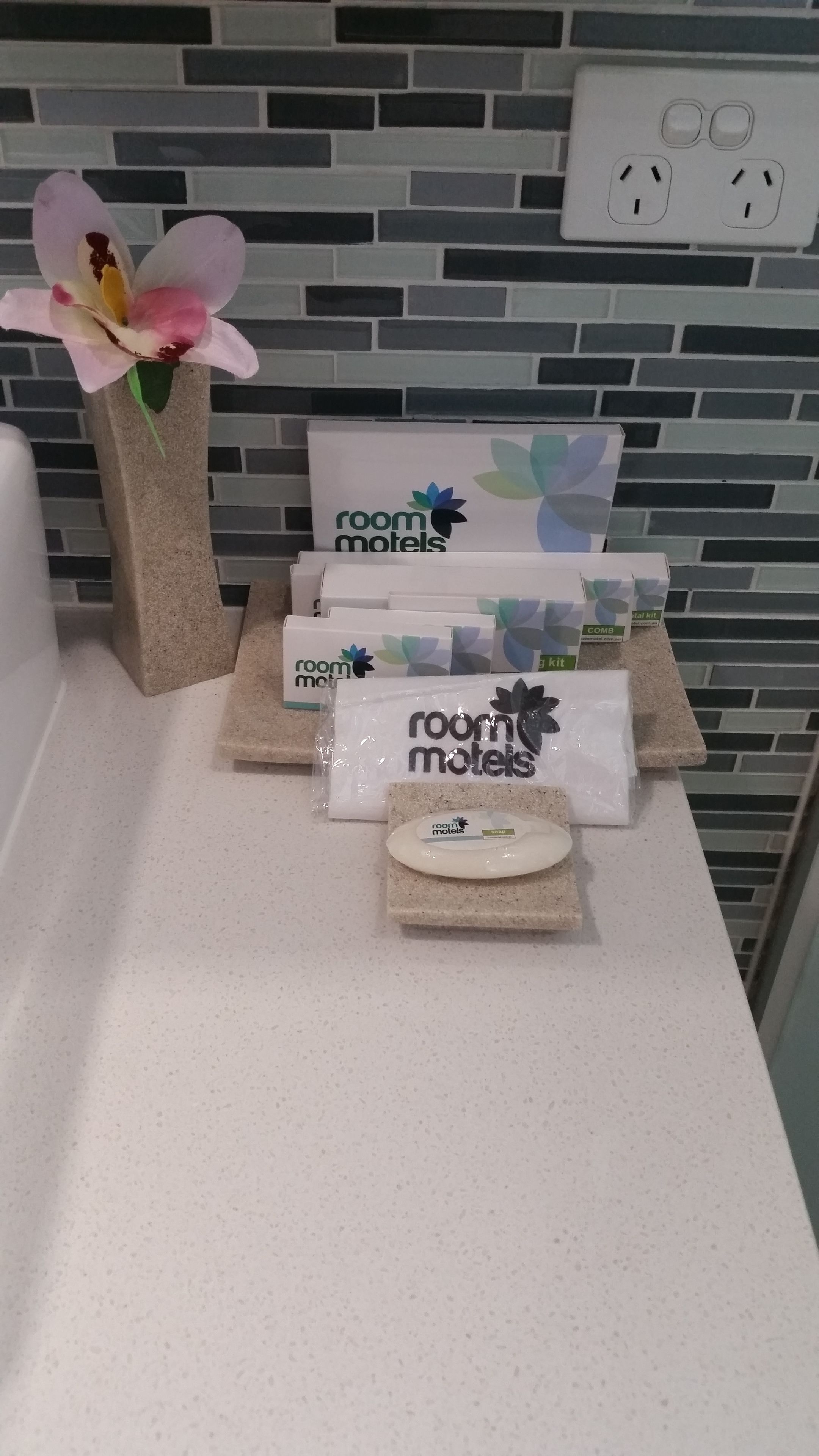 Habitación de hotel con un mostrador blanco que exhibe artículos esenciales como un peine, un espejo redondo pequeño y una bolsa de plástico con artículos de aseo. También se observa un florero con una flor rosa.