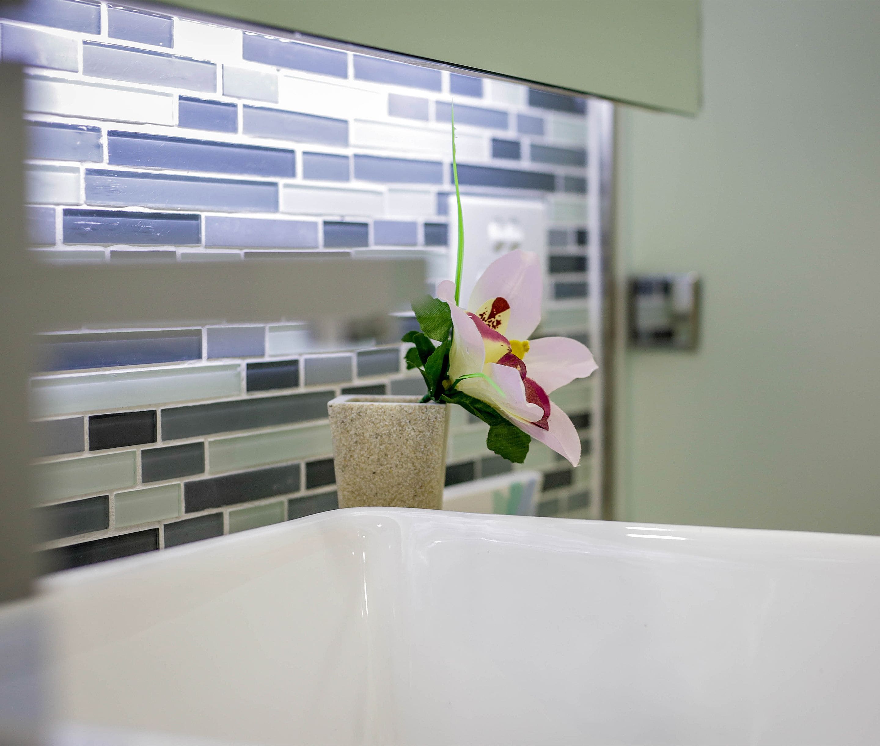 A bathroom with a white bathtub, a flower in a stone vase, and a tiled wall with a brick pattern.