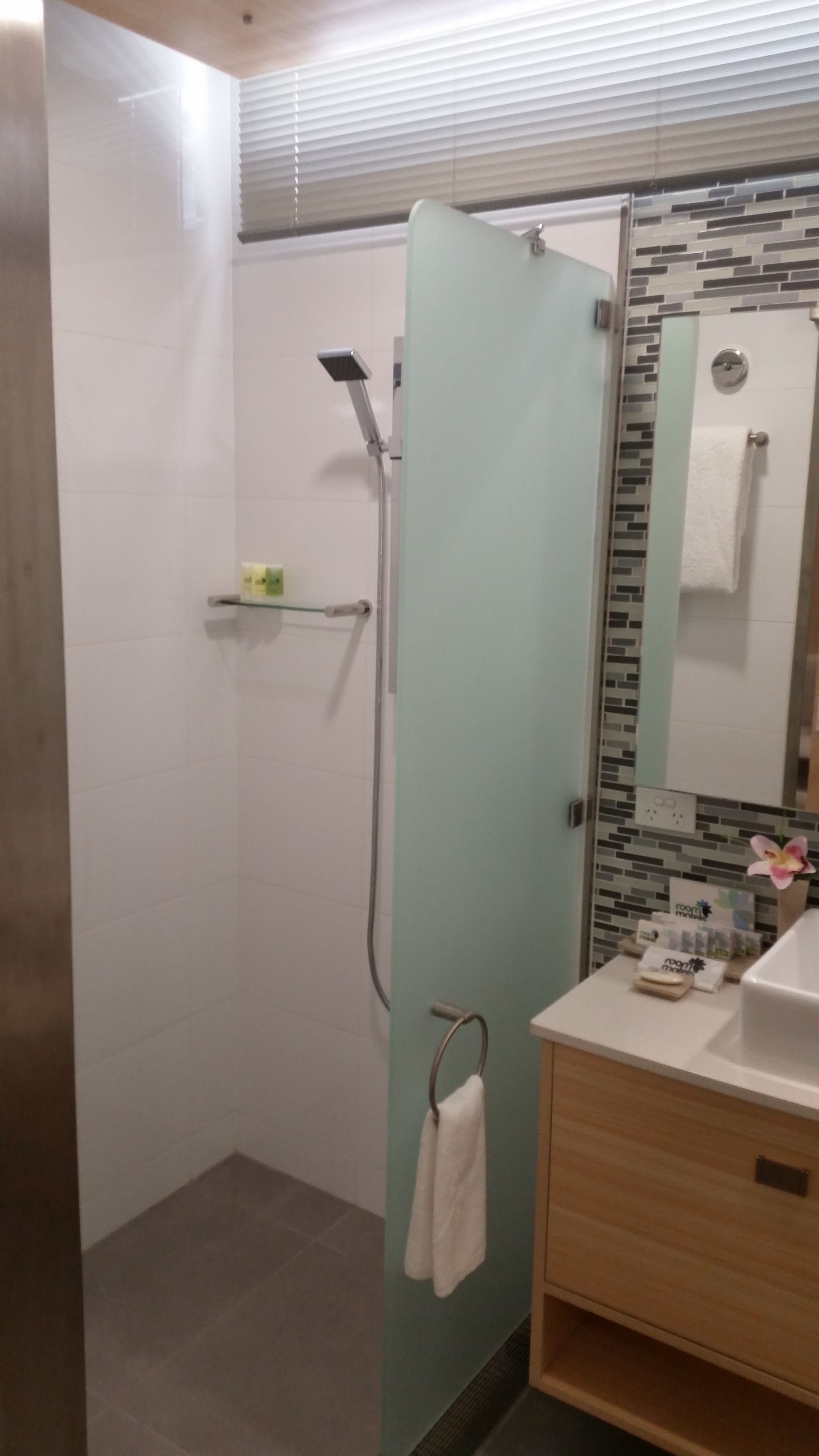 Et moderne badeværelse med glasvæg omkring bruseniche, et håndklædetørrestativ, en vask med toiletartikler og et spejl med et håndklæde hængende på.