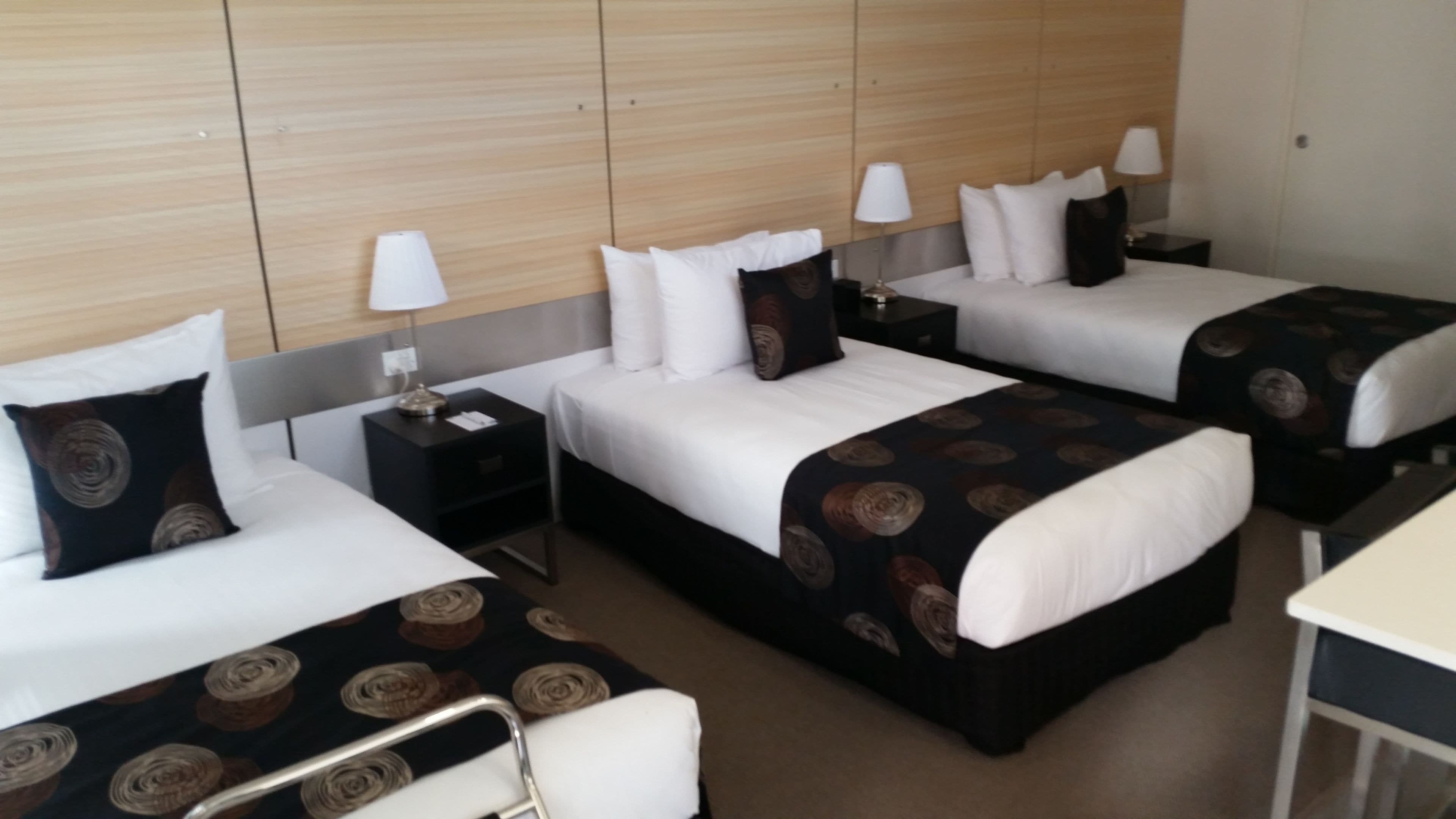 Quarto de hotel com três camas, cada uma com roupa de cama branca e colchas estampadas em tons escuros. Há mesas de cabeceira com abajures, uma pequena escrivaninha e uma cadeira.