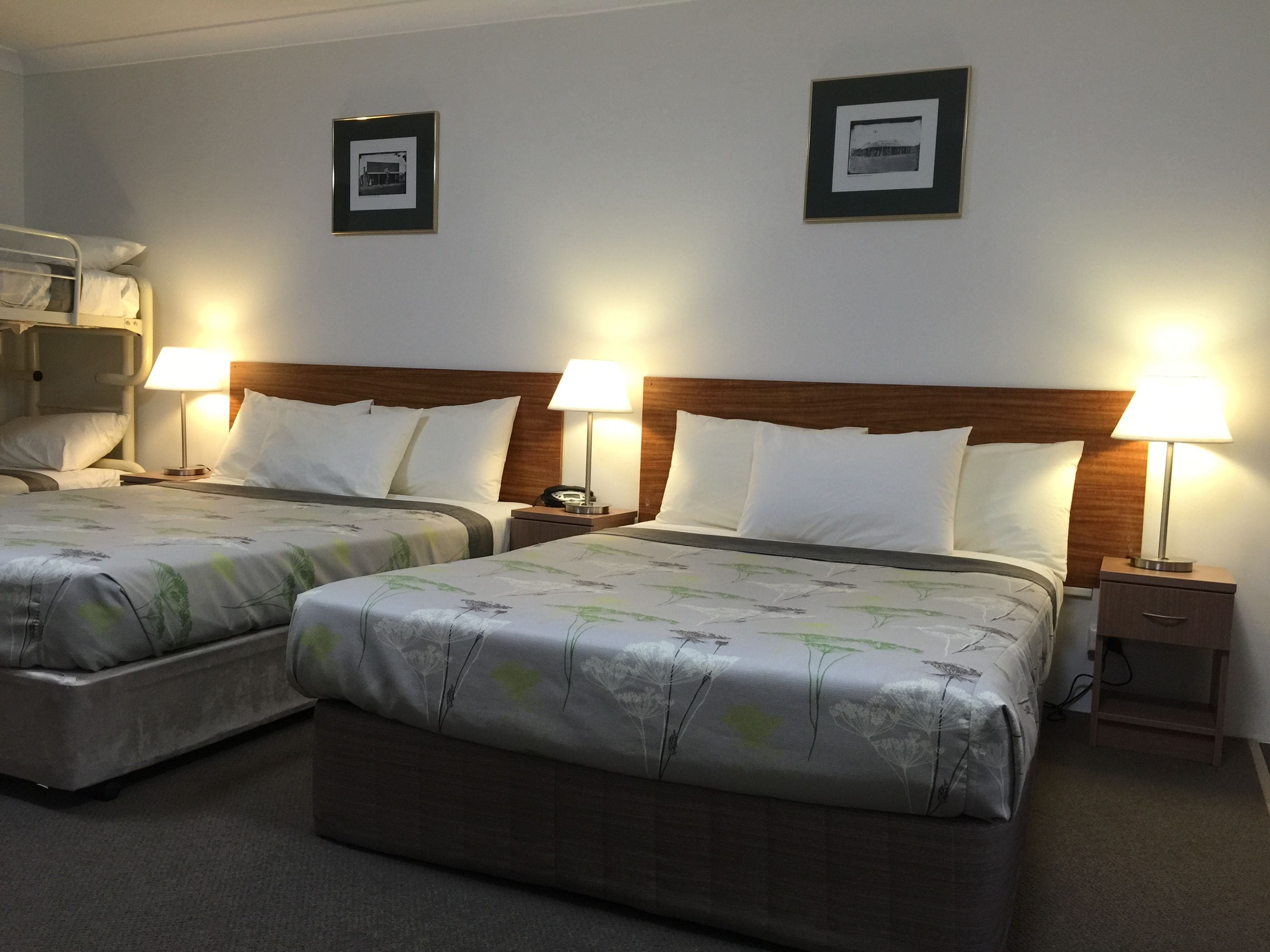 A hotel room with two beds, each with a wooden headboard, white bedding, and bedside lamps. There are framed pictures on the wall above each bed.
