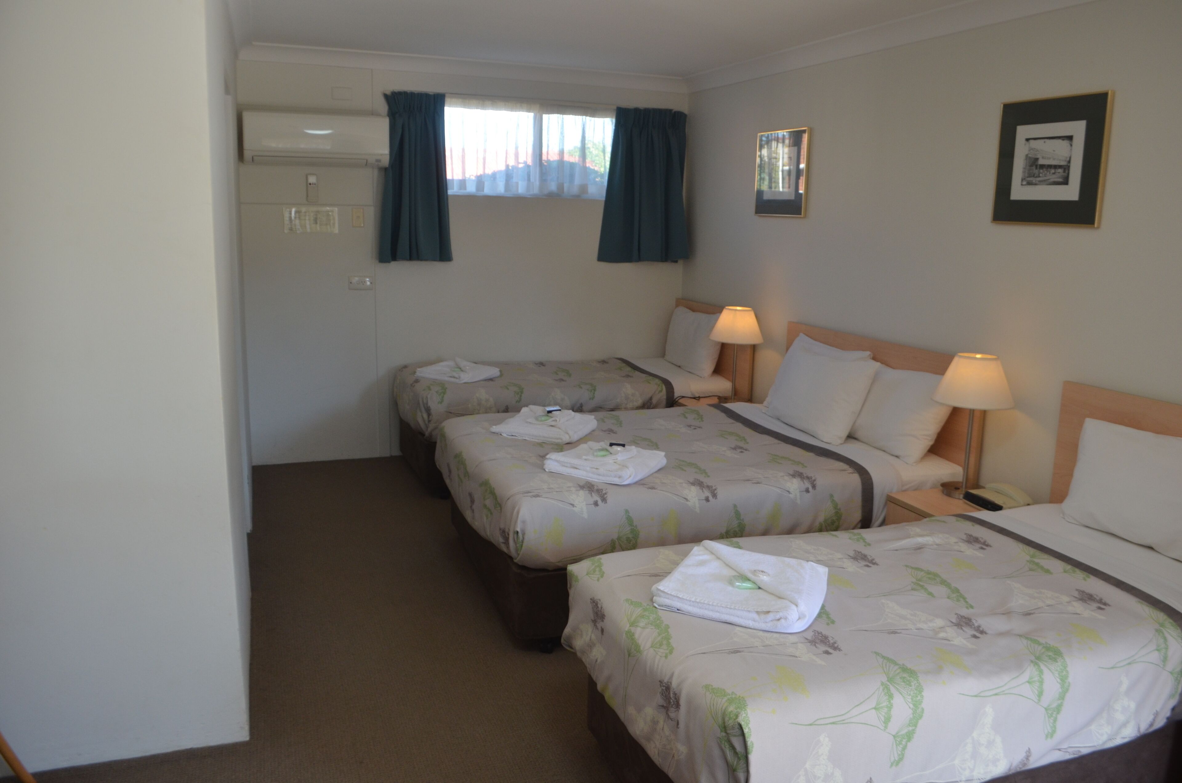Ein Hotelzimmer mit zwei Einzelbetten, einem kleinen Tisch, einer Lampe, einem Fenster mit Vorhängen und einer Wand-Klimaanlage.