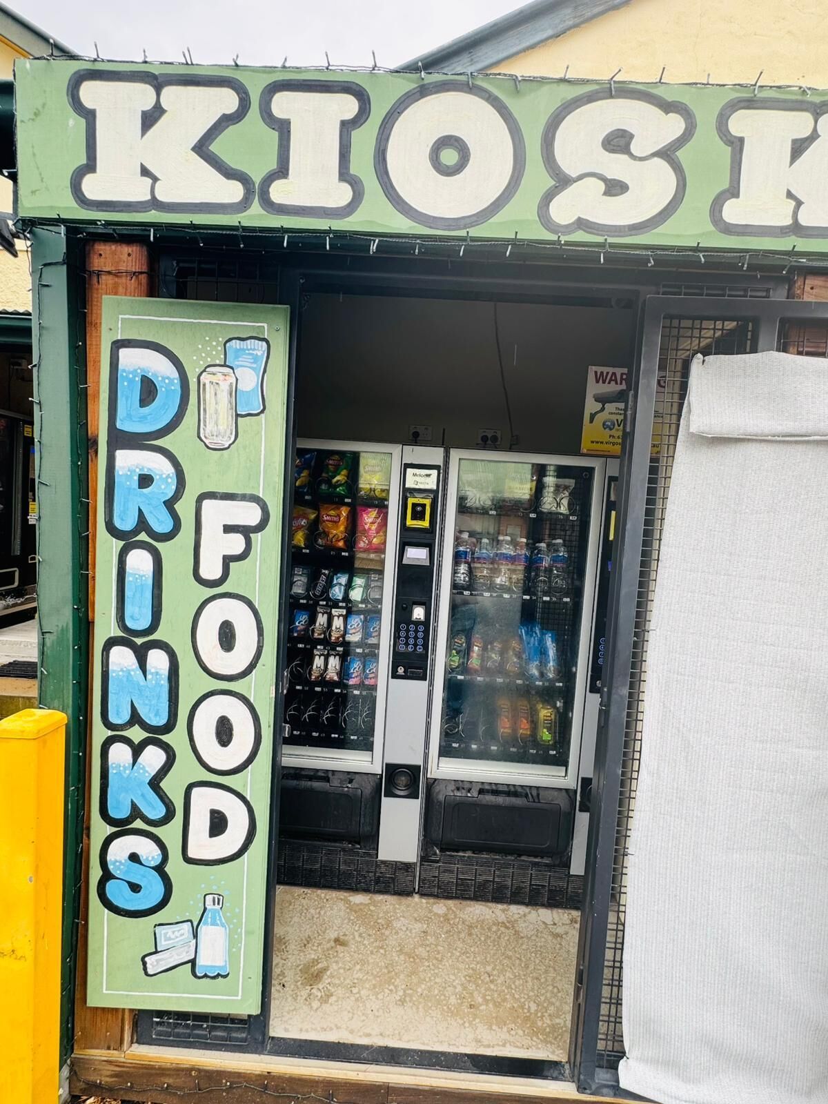 Ein Kiosk mit einem Schild, auf dem ‚DRINKS FOOD‘ steht, und Verkaufsautomaten im Inneren.