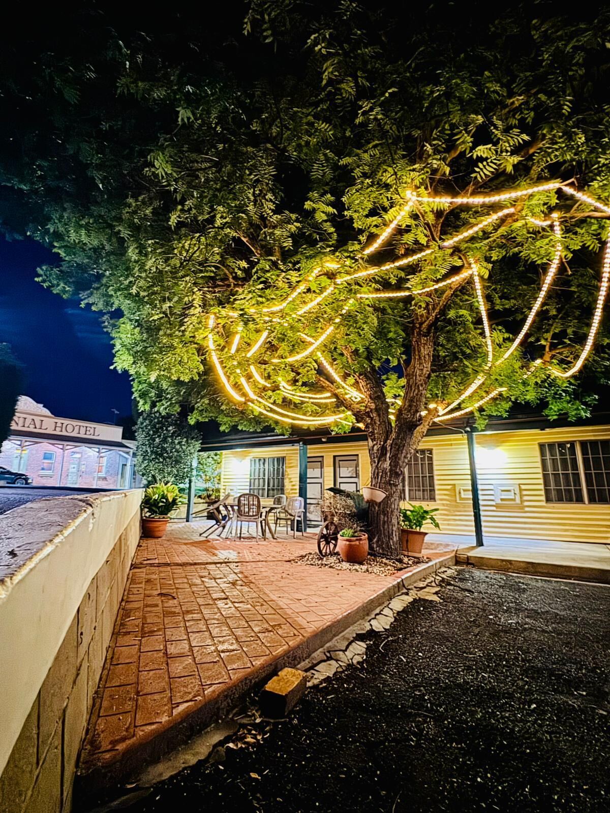 A tree with string lights, a brick pathway, outdoor seating, and a building labeled "HOTEL" in the background.
