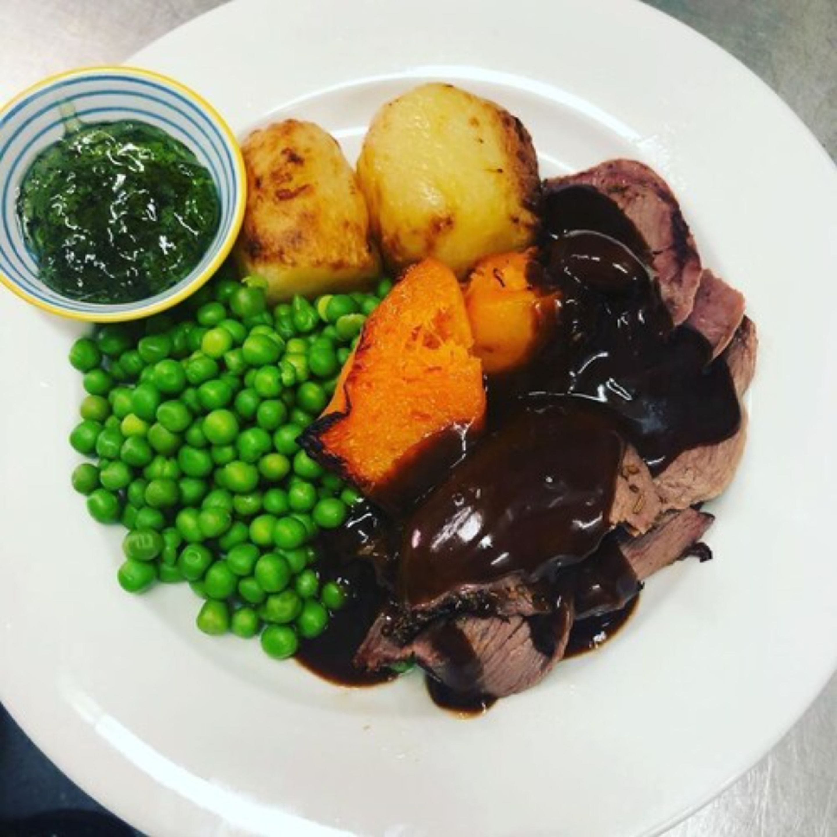 Ein Teller mit Braten, Kartoffelpüree, Erbsen und einer Soßenspender.
