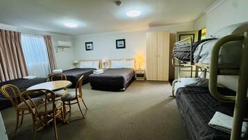 Habitación de hotel con dos camas individuales, una litera, una mesa de comedor con sillas, un armario y un escritorio con lámpara.