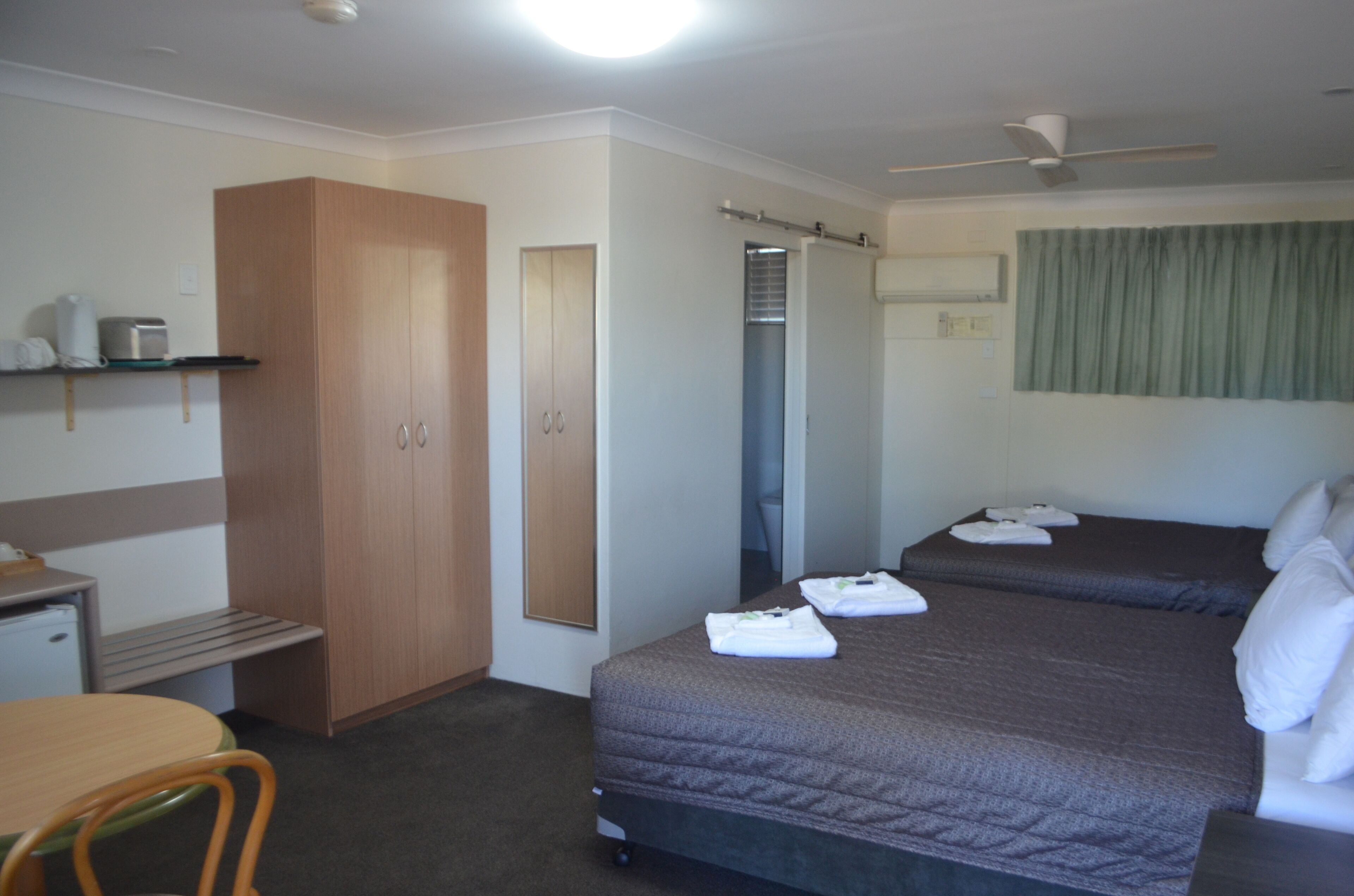 A hotel room with two beds, a desk with a chair, a wardrobe, and a shower area.