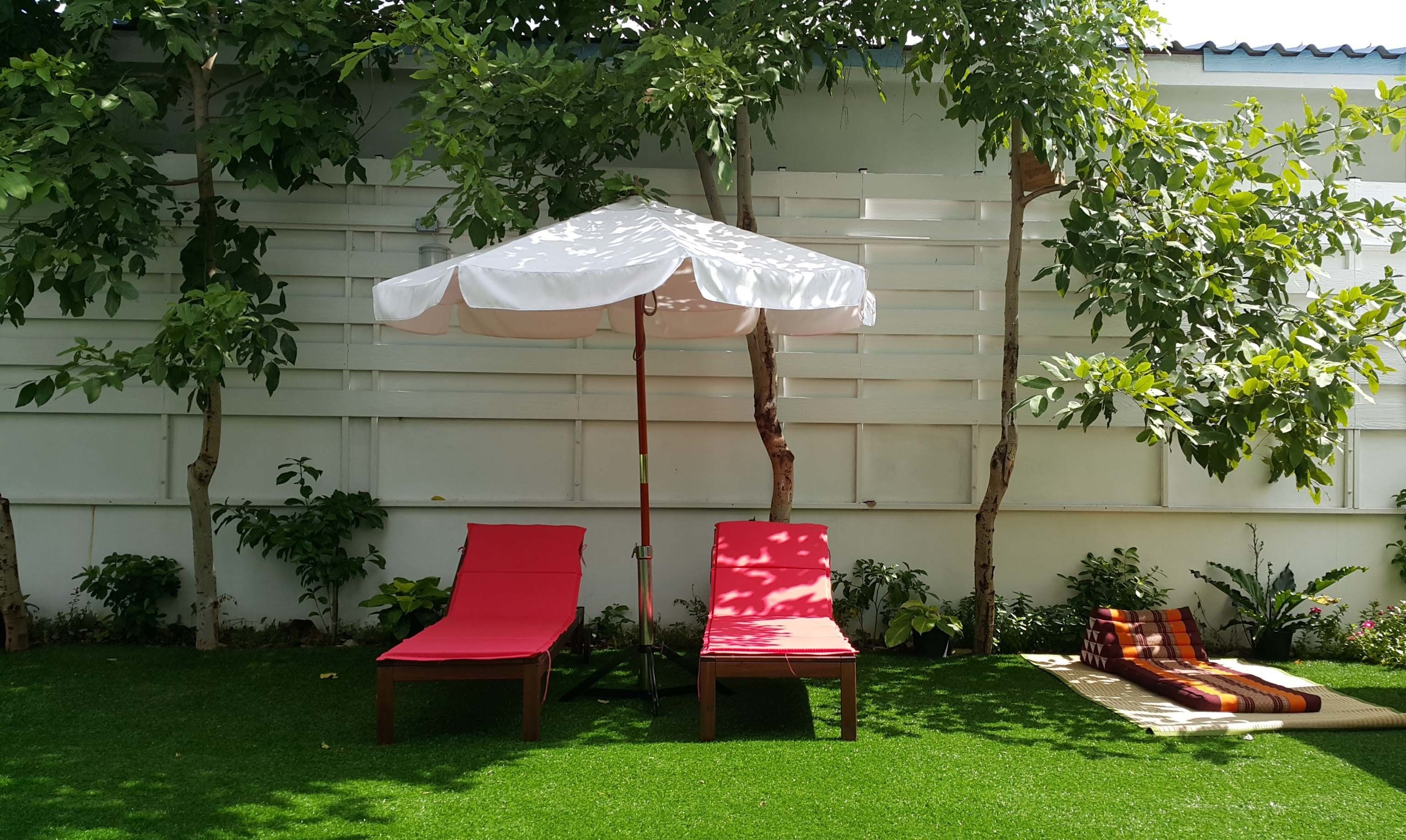 Zwei rote Liegestühle unter einem weißen Sonnenschirm in einem Garten mit weißem Zaun und Grünpflanzen.