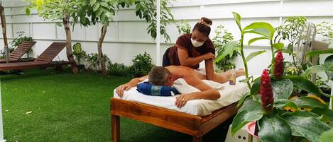 A wooden bed in a garden setting with a person lying on it, another person attending to them, and various plants around.