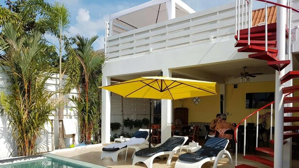 A pool area with lounge chairs and a yellow umbrella, surrounded by tropical plants. - Barefeet Naturist Resort (Bangkok)
