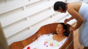 A wooden tub filled with water and flower petals, with a person standing beside it. - Barefeet Naturist Resort (Bangkok)