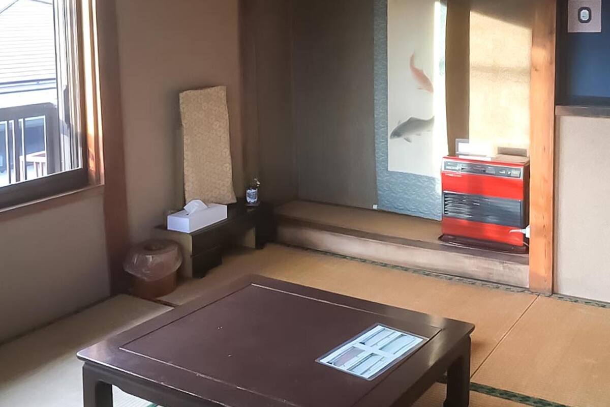 Une pièce avec un sol recouvert de tatamis, une table en bois avec un petit présentoir, une petite poubelle et un meuble rouge équipé d’un distributeur d’eau.