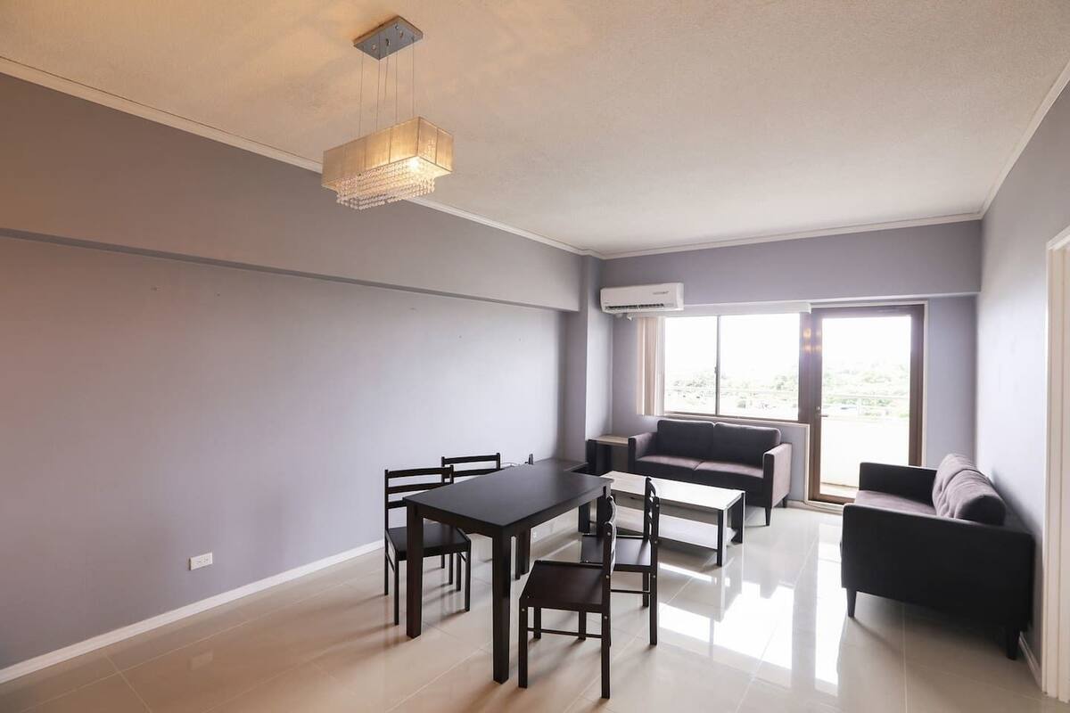 A modern living room with a dining table, chairs, a sofa, and a large window providing natural light.