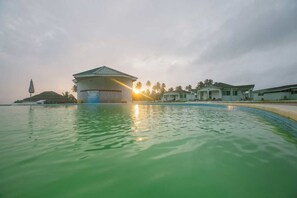 A swimming pool with a building in the background, surrounded by palm trees and a cloudy sky. - Karela Beach Resort (Beyin)