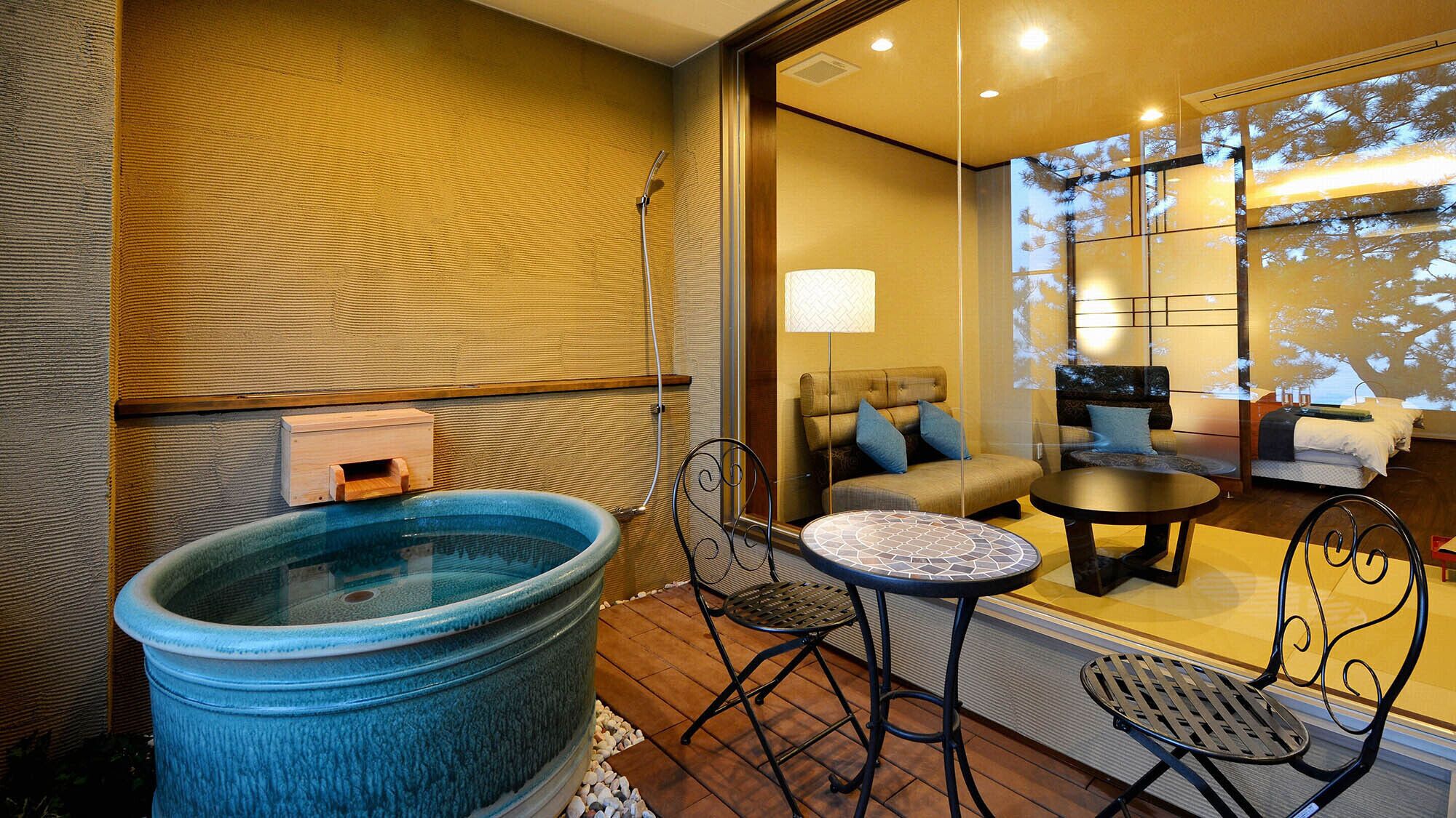 A room with a large blue hot tub, a small round table, two chairs, a sofa, and a bed with a view of trees through a window.