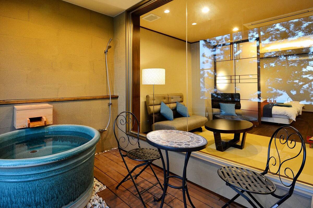 A room with a large blue hot tub, a small round table, two chairs, a sofa, and a bed with a view of trees through a window.