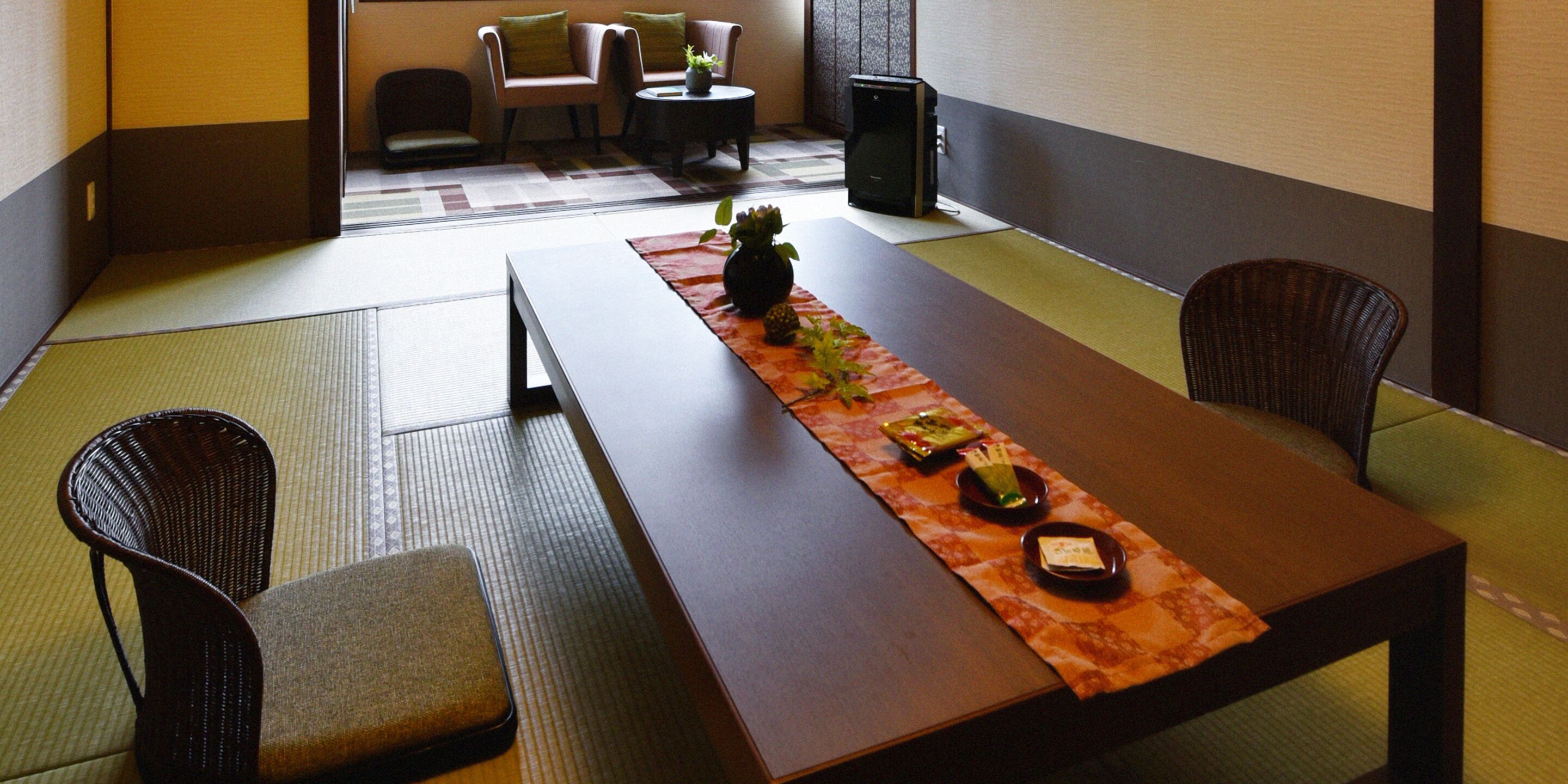 A traditional Japanese room with tatami flooring, a low table with a placemat, and a small plant. There are two chairs and a sofa with cushions.