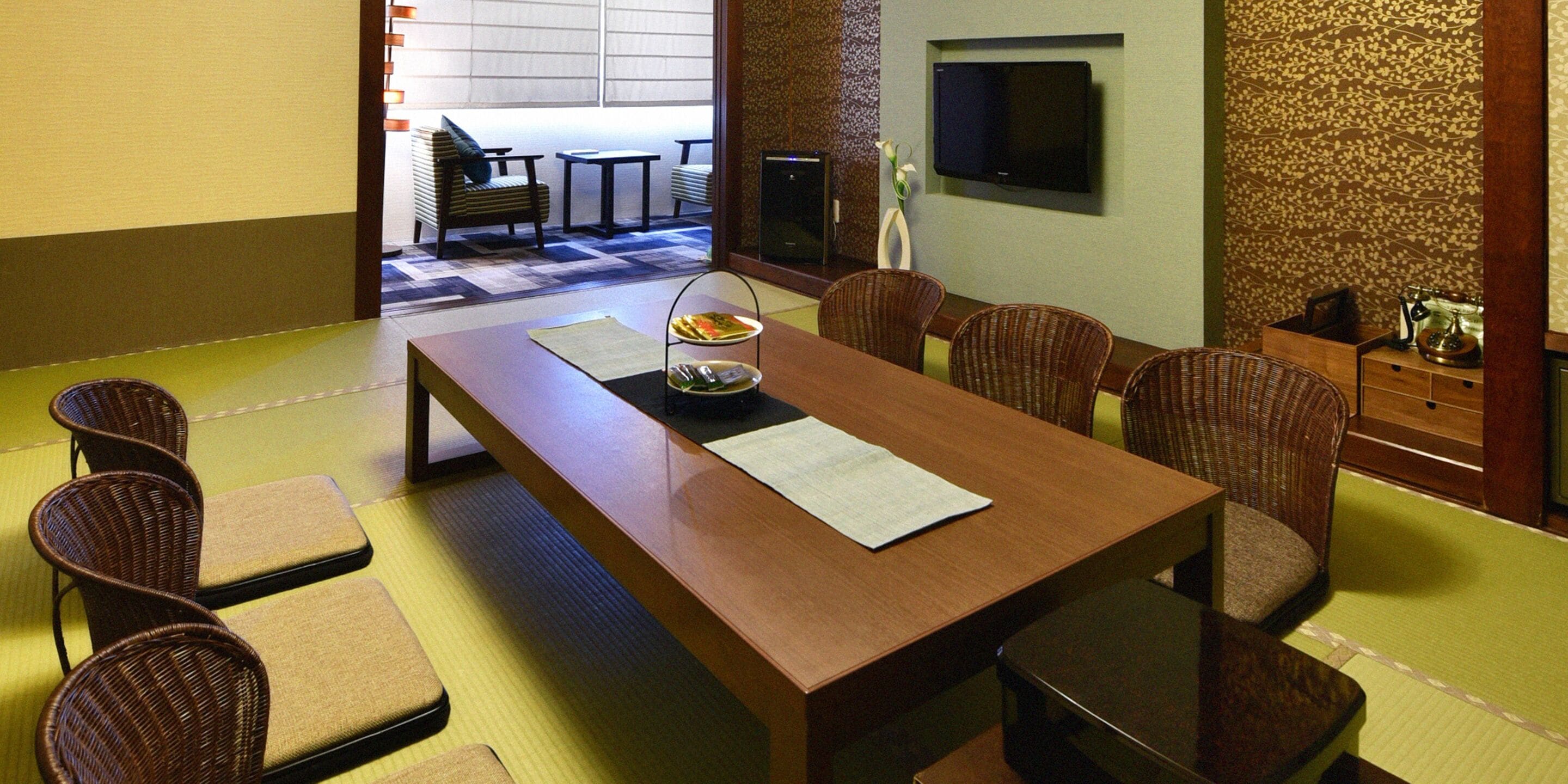 A dining area with a long wooden table, several wicker chairs, a small side table with a vase, and a television mounted on the wall.