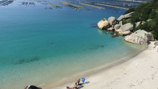 Una playa costera con agua cristalina, afloramientos rocosos y una vista distante de un puerto pesquero.
