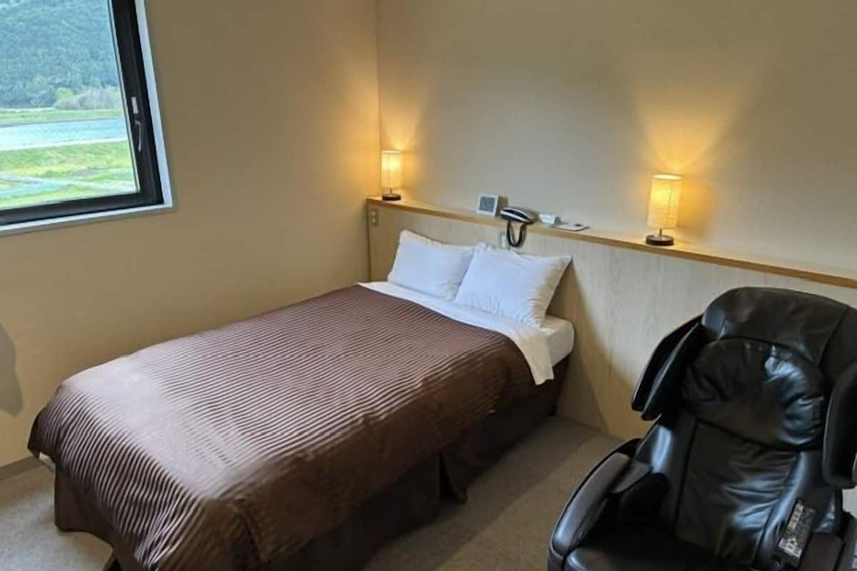 Uma cama de solteiro com colcha marrom, duas luminárias de cabeceira, encosto da cama, telefone e janela com vista para o verde.