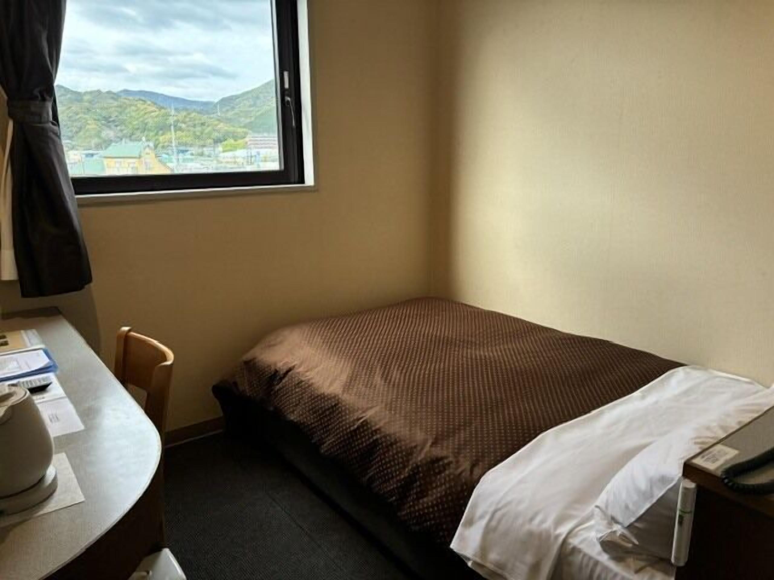 Une petite chambre d’hôtel avec un lit simple, un bureau avec une chaise et une fenêtre donnant sur une montagne et des bâtiments.