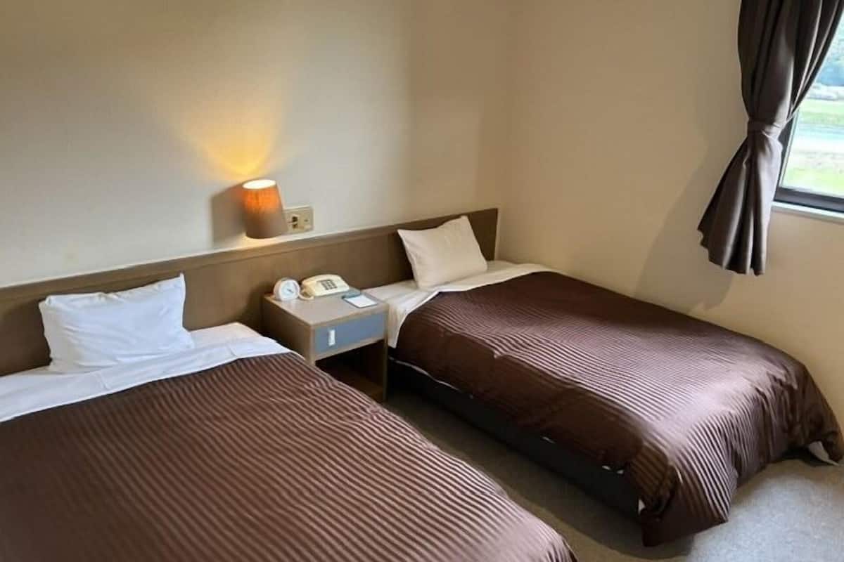 Quarto de hotel com duas camas, uma mesa de cabeceira com abajur, telefone e janela com cortinas.